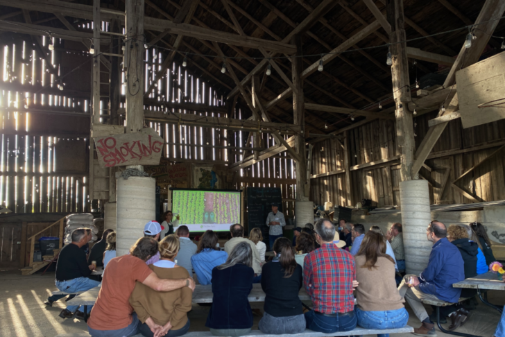 A group of people in a barn watching a TV screen displaying a farmer’s field